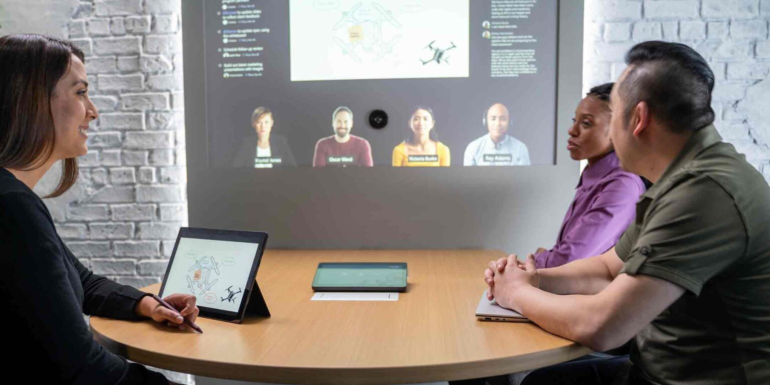 Business people sitting at desk with Microsoft Teams on a large screen near them