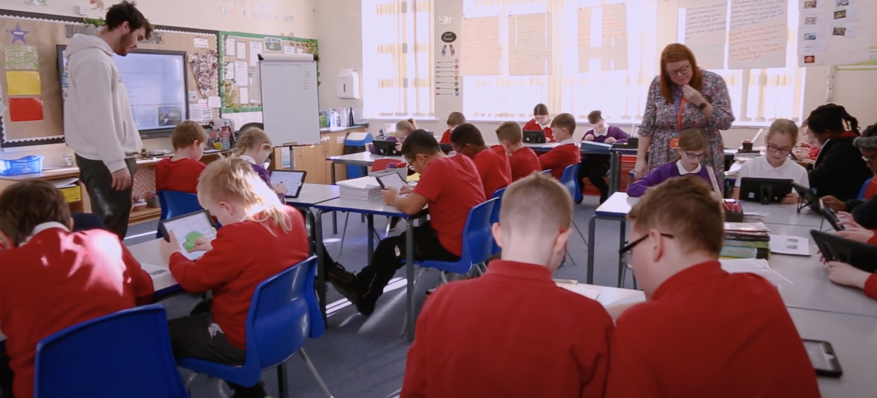 Schoolchildren in red school uniform using tablets in a classroom as a teacher supervises